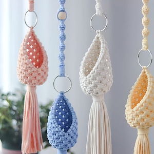 May include: Four macrame plant hangers with different coloured pots. The pots are light pink, white, light blue, and light yellow. Each pot has a tassel hanging from the bottom.