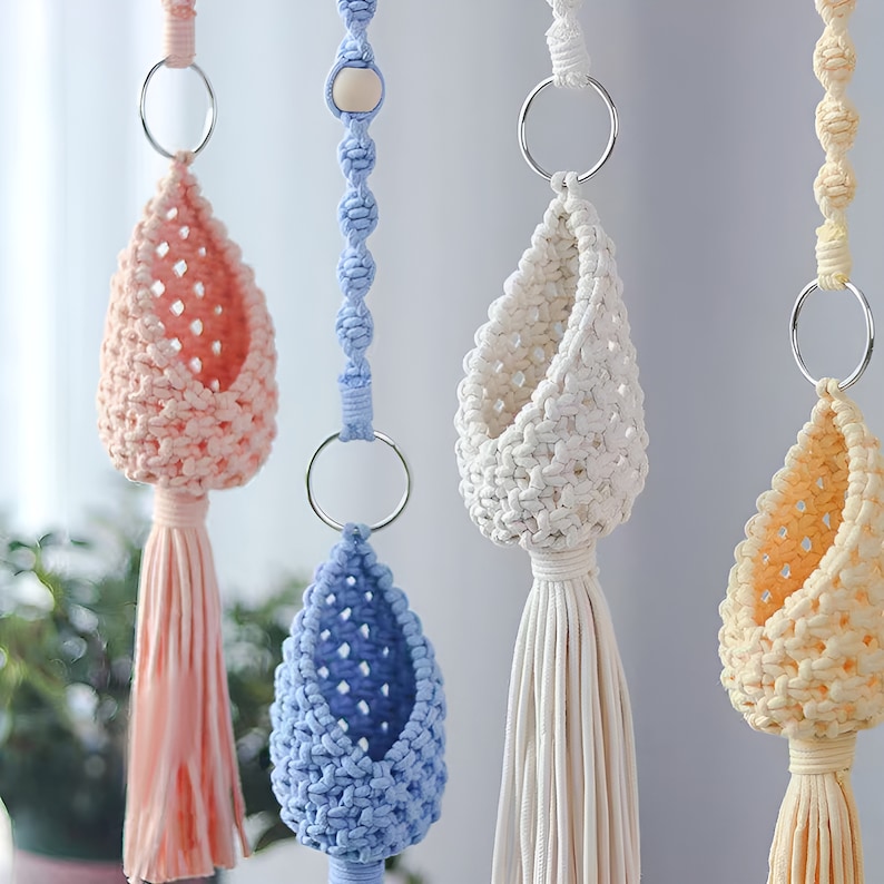May include: Four macrame plant hangers with different colored pots. The pots are light pink, white, light blue, and light yellow. Each pot has a tassel hanging from the bottom.