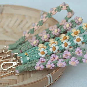 May include: A set of colorful macrame keychains with daisy flower designs. The keychains are made with green, pink, and yellow string and feature gold metal clasps.