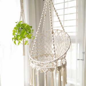 Op de afbeelding: Een witte macramé hangstoel met houten kralen en kwastjes. De stoel hangt aan een wit plafond.