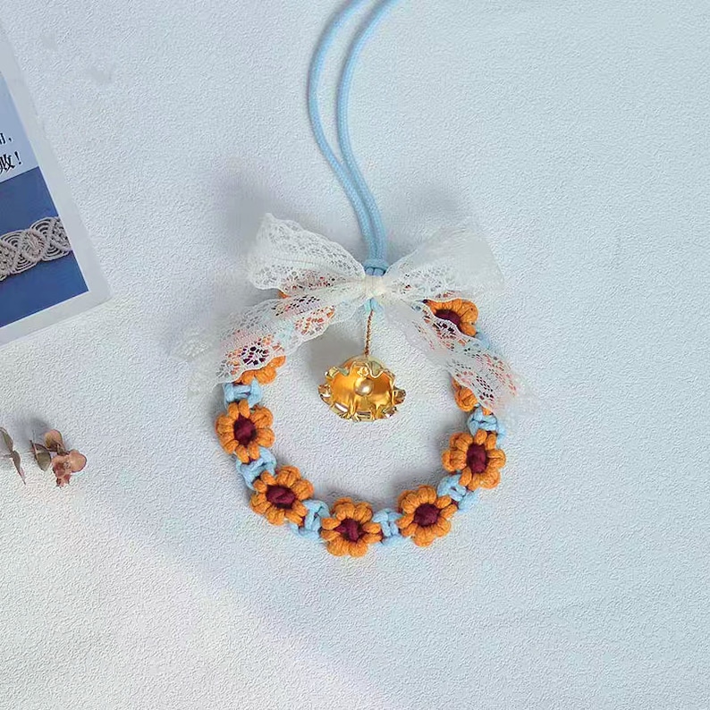 K&ouml;nnte beinhalten: Ein blauer und orangefarbener Makramee-Kranz mit einer goldenen Glocke und einer wei&szlig;en Schleife aus Spitze. Der Kranz h&auml;ngt an einer blauen Schnur.