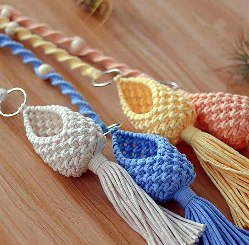 May include: Three macrame plant hangers with tassels in cream, light blue, and yellow. The hangers are made with a knotted rope and have a metal ring at the top for hanging.