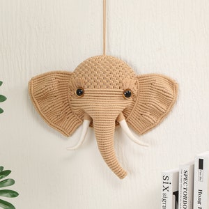 以下が含まれることがあります： 白い壁に掛けられた、ベージュの毛糸で編まれた象の壁飾り。白い毛糸の牙と黒い目がある。
