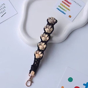 K&ouml;nnte beinhalten: Ein schwarzer und beiger geflochtener Schl&uuml;sselanh&auml;nger mit einem goldenen Metallverschluss. Der Schl&uuml;sselanh&auml;nger ist aus gewebtem Stoff gefertigt und hat eine Reihe kleiner, runder Perlen.