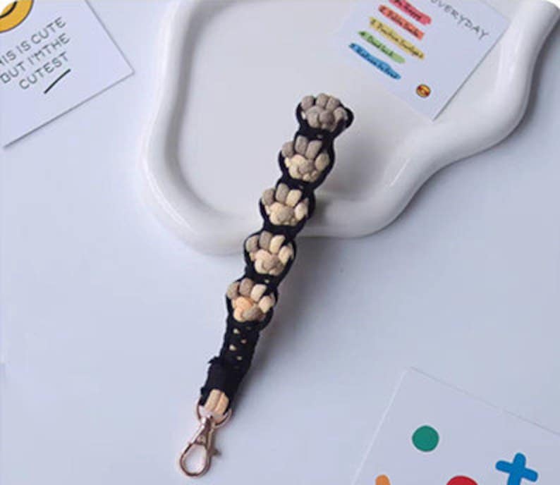 K&ouml;nnte beinhalten: Ein schwarzer und beiger geflochtener Schl&uuml;sselanh&auml;nger mit einem goldenen Metallverschluss. Der Schl&uuml;sselanh&auml;nger ist aus gewebtem Stoff gefertigt und hat eine Reihe kleiner, runder Perlen.