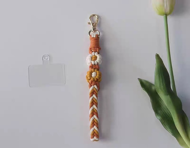 Pu&ograve; includere: Un portachiavi intrecciato bianco e arancione con una chiusura dorata e tre accenti di fiori bianchi e gialli.