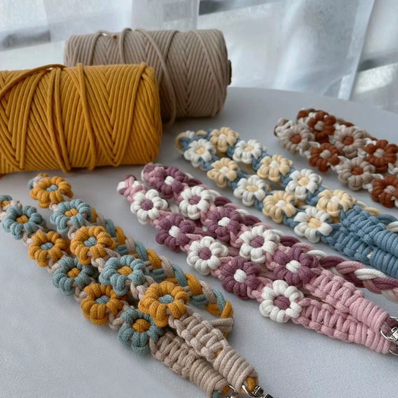 May include: A collection of handmade macrame cord bracelets with daisy flower designs in various colors, including beige, yellow, blue, pink, and white. The cords are arranged in a row on a white surface.