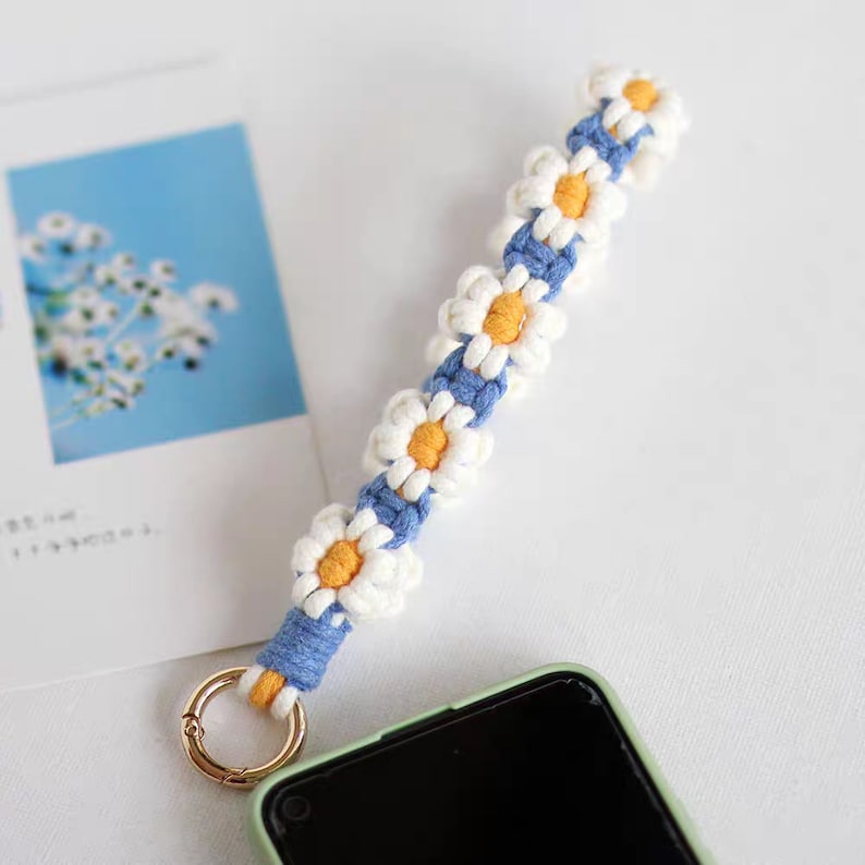 Puede incluir: Una correa para tel&eacute;fono blanca y azul con un dise&ntilde;o de margarita. La correa tiene un anillo de metal dorado en la parte superior.