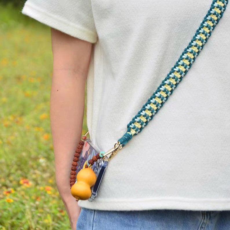May include: A teal, yellow, and white braided phone strap with a gold clasp. The strap is attached to a clear phone case with a brown gourd charm.