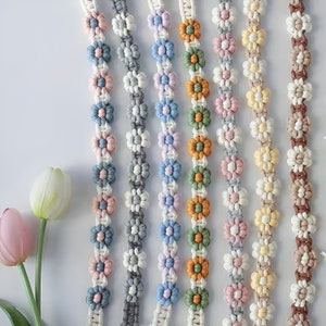 Op de afbeelding: Assortiment van kleurrijke gecrochete bloemtjesbies met verschillende kleuren, waaronder blauw, groen, geel, roze en bruin. De bies is gemaakt van katoenen garen en heeft een madeliefje-ontwerp.