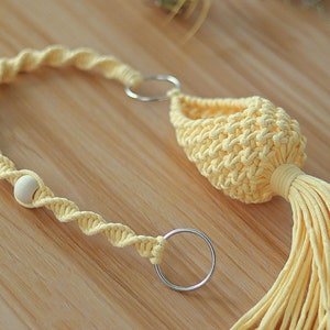 May include: A yellow macrame plant hanger with a wooden bead and two metal rings. The hanger has a tassel at the bottom.