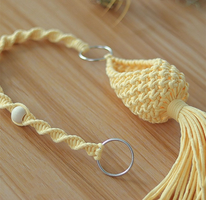 May include: A yellow macrame plant hanger with a wooden bead and two metal rings. The hanger has a tassel at the bottom.
