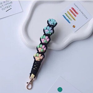 K&ouml;nnte beinhalten: Ein schwarzer und mehrfarbiger geflochtener Schl&uuml;sselanh&auml;nger mit einem goldenen Metallverschluss. Der Schl&uuml;sselanh&auml;nger besteht aus einem weichen, gewebten Material und verf&uuml;gt &uuml;ber eine Reihe von bunten Knoten.