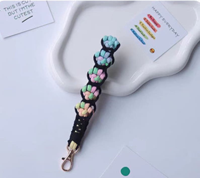 K&ouml;nnte beinhalten: Ein schwarzer und mehrfarbiger geflochtener Schl&uuml;sselanh&auml;nger mit einem goldenen Metallverschluss. Der Schl&uuml;sselanh&auml;nger besteht aus einem weichen, gewebten Material und verf&uuml;gt &uuml;ber eine Reihe von bunten Knoten.