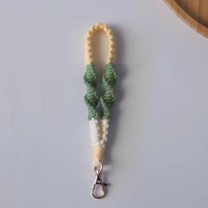 Pode incluir: Um chaveiro de croch&ecirc; amarelo, branco e verde com um fecho de metal prateado.