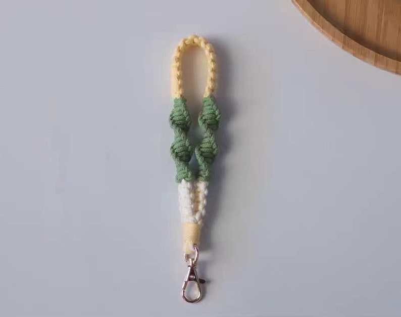 Pode incluir: Um chaveiro de croch&ecirc; amarelo, branco e verde com um fecho de metal prateado.