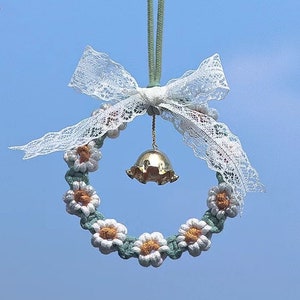 K&ouml;nnte beinhalten: Ein wei&szlig;er H&auml;kelkranz mit einer goldenen Glocke, die daran h&auml;ngt. Der Kranz ist mit wei&szlig;en und gelben Blumen und einer wei&szlig;en Schleife aus Spitze verziert.