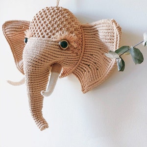 Puede incluir: Una cabeza de elefante de macram&eacute; beige con colmillos de hilo blanco y ojos marrones.
