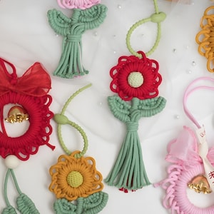 May include: A collection of handmade macrame flower ornaments in various colors, including pink, red, and yellow. Each flower features a detailed design with green stems and leaves. Some ornaments include decorative bows and bells.