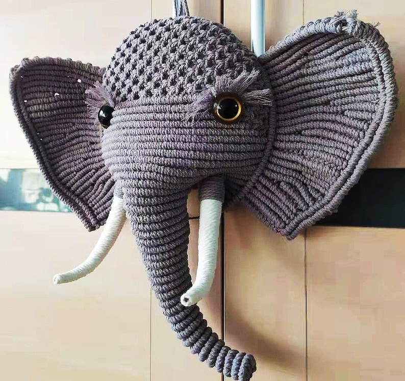 Puede incluir: Una cabeza de elefante de crochet gris con colmillos de hilo blanco y grandes ojos negros.