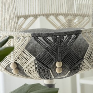 Op de afbeelding: Een witte en grijze macramé kattenhangmat met een grijs stoffen kussen. De hangmat hangt aan het plafond en heeft houten kralen aan de randen.