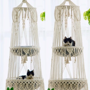 Op de afbeelding: Twee witte macramé kattenhangmatten met grijze kussens. Elke hangmat heeft twee niveaus en hangt aan het plafond. Een zwart-witte kat zit op het bovenste niveau van de ene hangmat, en een andere zwart-witte kat ligt op het onderste niveau van de andere hangmat.