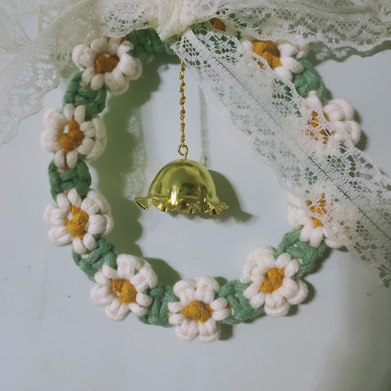 K&ouml;nnte beinhalten: Ein wei&szlig;er und gr&uuml;ner geh&auml;kelter Kranz mit einer goldenen Glocke, die daran h&auml;ngt. Der Kranz ist mit wei&szlig;en Blumen verziert und hat eine Schleife aus Spitze.