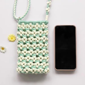 May include: A mint green and white macrame crossbody bag featuring a repeating flower pattern, a braided strap, and a rectangular body. A black smartphone is positioned next to the bag to provide a visual scale for its size.