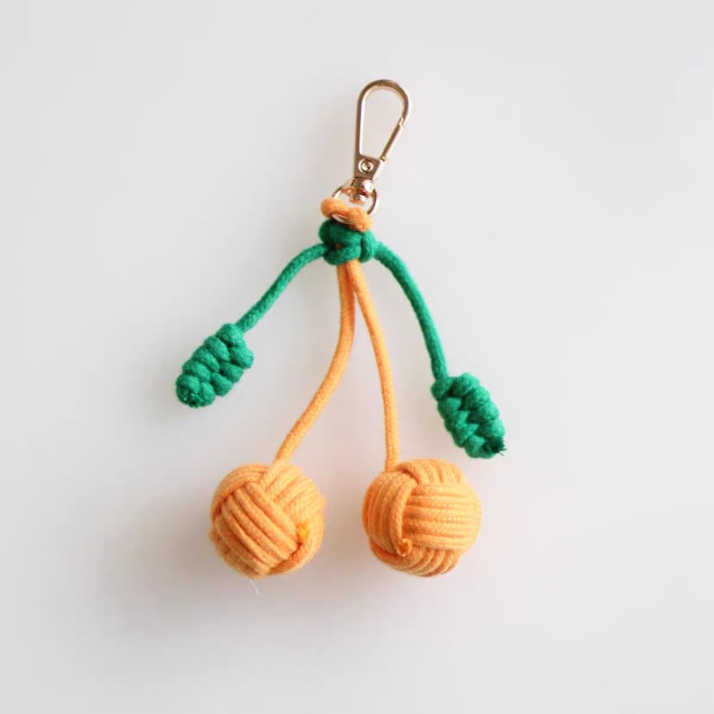 Op de afbeelding: Een sleutelhanger met twee oranje geknoopte kersen en groene geknoopte bladeren. De sleutelhanger heeft een gouden metalen sluiting.