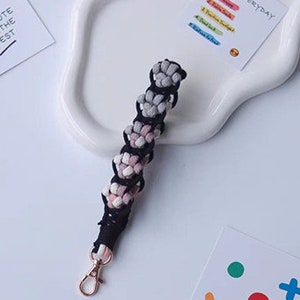 K&ouml;nnte beinhalten: Ein schwarzer und wei&szlig;er geflochtener Schl&uuml;sselanh&auml;nger mit einem rosa und grauen Ombre-Effekt. Der Schl&uuml;sselanh&auml;nger hat einen goldenen Schl&uuml;sselring aus Metall.