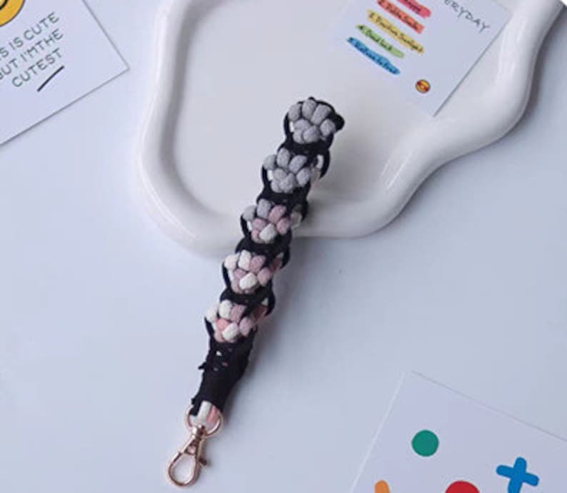 K&ouml;nnte beinhalten: Ein schwarzer und wei&szlig;er geflochtener Schl&uuml;sselanh&auml;nger mit einem rosa und grauen Ombre-Effekt. Der Schl&uuml;sselanh&auml;nger hat einen goldenen Schl&uuml;sselring aus Metall.