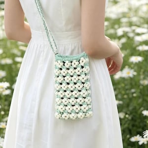 May include: A rectangular crossbody bag crafted from crocheted yarn in a repeating floral pattern. The bag features a light mint green and cream color scheme with a braided strap, designed to hold small personal items securely.
