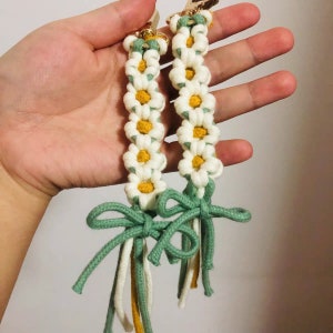 May include: Two white and green macrame keychains with daisy flower designs and green bows.