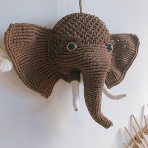 Puede incluir: Una cabeza de elefante de crochet marrón con colmillos de hilo blanco y ojos de botón negros.