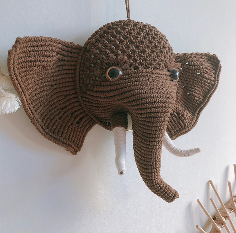 Puede incluir: Una cabeza de elefante de crochet marr&oacute;n con colmillos de hilo blanco y ojos de bot&oacute;n negros.