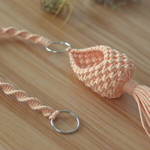 May include: A peach-colored macrame plant hanger with a tassel. The hanger has two silver metal rings for hanging.