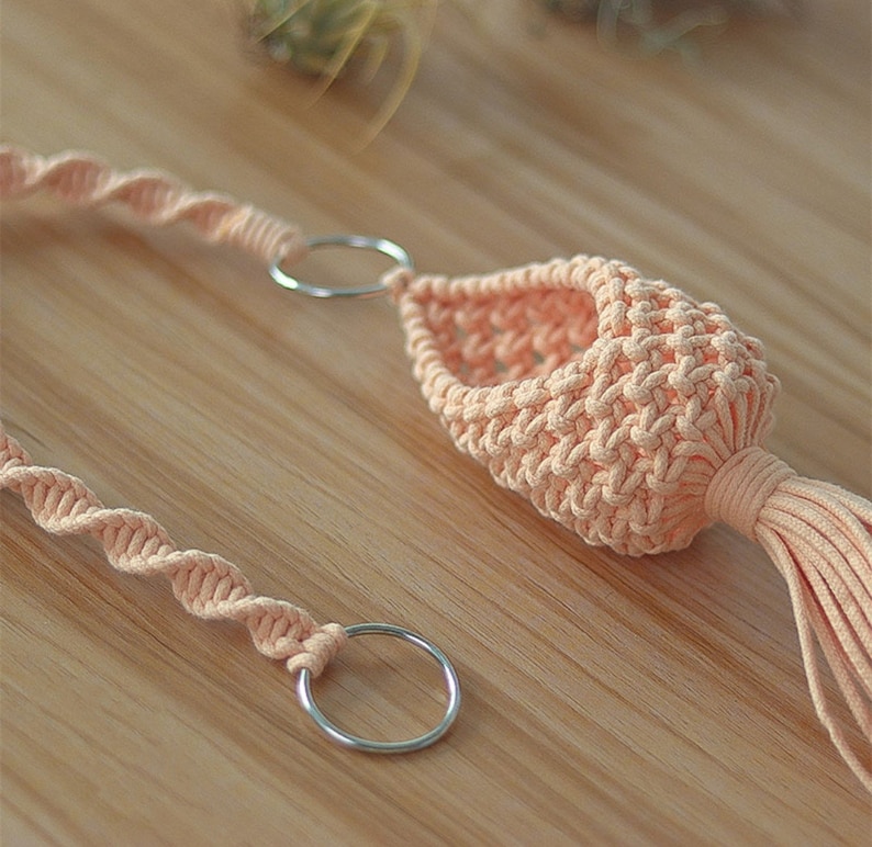 May include: A peach-colored macrame plant hanger with a tassel. The hanger has two silver metal rings for hanging.