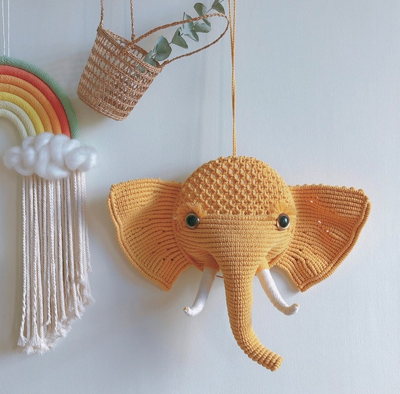 Puede incluir: Una cabeza de elefante de crochet amarilla colgada en la pared con ojos negros. El elefante est&aacute; colgado de una cuerda y tiene una superficie texturizada.