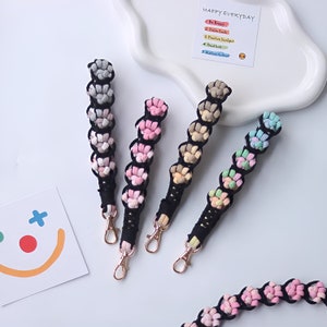 Op de afbeelding: Vijf kleurrijke sleutelhangers met een zwarte gevlochten band en een gouden metalen sluiting. Elke sleutelhanger is gemaakt van kleine pompoenen in verschillende kleuren, waaronder roze, grijs, beige, lichtblauw en groen. De sleutelhangers zijn in een rij op een wit oppervlak gerangschikt. De tekst "HAPPY EVERYDAY" staat op een witte kaart op de achtergrond.