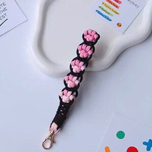 K&ouml;nnte beinhalten: Ein rosa-schwarzer geflochtener Schl&uuml;sselanh&auml;nger mit einem goldenen Metallverschluss. Der Schl&uuml;sselanh&auml;nger ist aus Garn gefertigt und hat eine Reihe von rosa Bommeln.