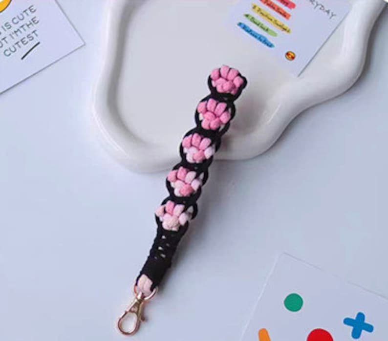 K&ouml;nnte beinhalten: Ein rosa-schwarzer geflochtener Schl&uuml;sselanh&auml;nger mit einem goldenen Metallverschluss. Der Schl&uuml;sselanh&auml;nger ist aus Garn gefertigt und hat eine Reihe von rosa Bommeln.