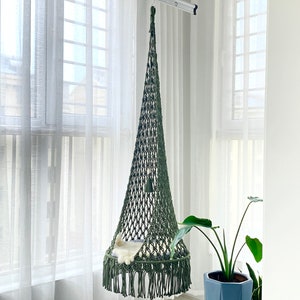 May include: A green macrame cat hammock hanging from a ceiling hook. The hammock is made of thick rope and has a fringe around the bottom. A white cat is sleeping inside the hammock.