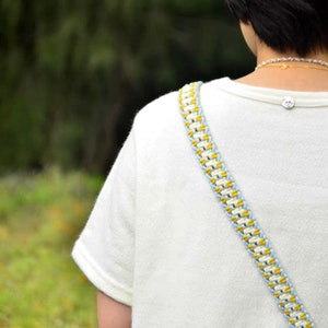May include: A white t-shirt with a blue and yellow braided strap. The strap is worn over the shoulder and is a woven design.