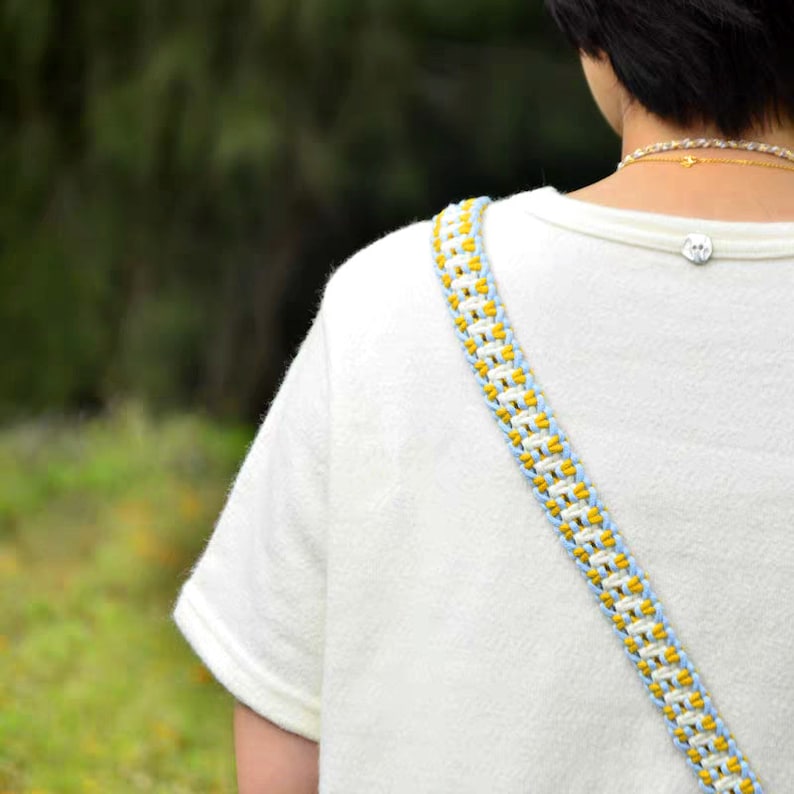 May include: A white t-shirt with a blue and yellow braided strap. The strap is worn over the shoulder and is a woven design.