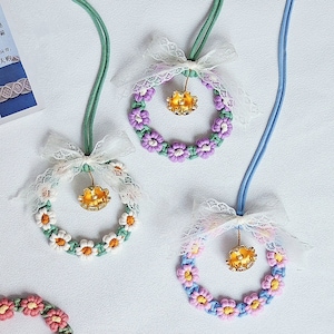 Könnte beinhalten: Drei Halsketten mit Blumenkränzen und einem goldenen Glöckchen-Anhänger. Jede Halskette hat eine weiße Schleife aus Spitze. Die Kränze sind aus buntem Garn gefertigt und haben ein Gänseblümchen-Design. Die Halsketten liegen auf einem weißen Hintergrund.