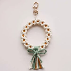 May include: White daisy flower keychain with a green and yellow tassel. The keychain has a gold metal clasp.