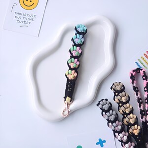 K&ouml;nnte beinhalten: Ein schwarz-wei&szlig;er Schl&uuml;sselanh&auml;nger mit einer Reihe von bunten, geh&auml;kelten Blumenperlen. Der Schl&uuml;sselanh&auml;nger hat einen goldenen Metallverschluss.