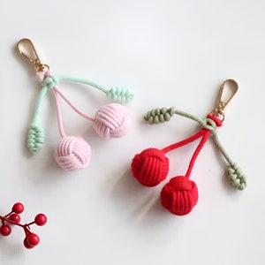 Op de afbeelding: Twee handgeknoopte kers-sleutelhangers. Een hanger is roze met groene bladeren en de andere is rood met groene bladeren.