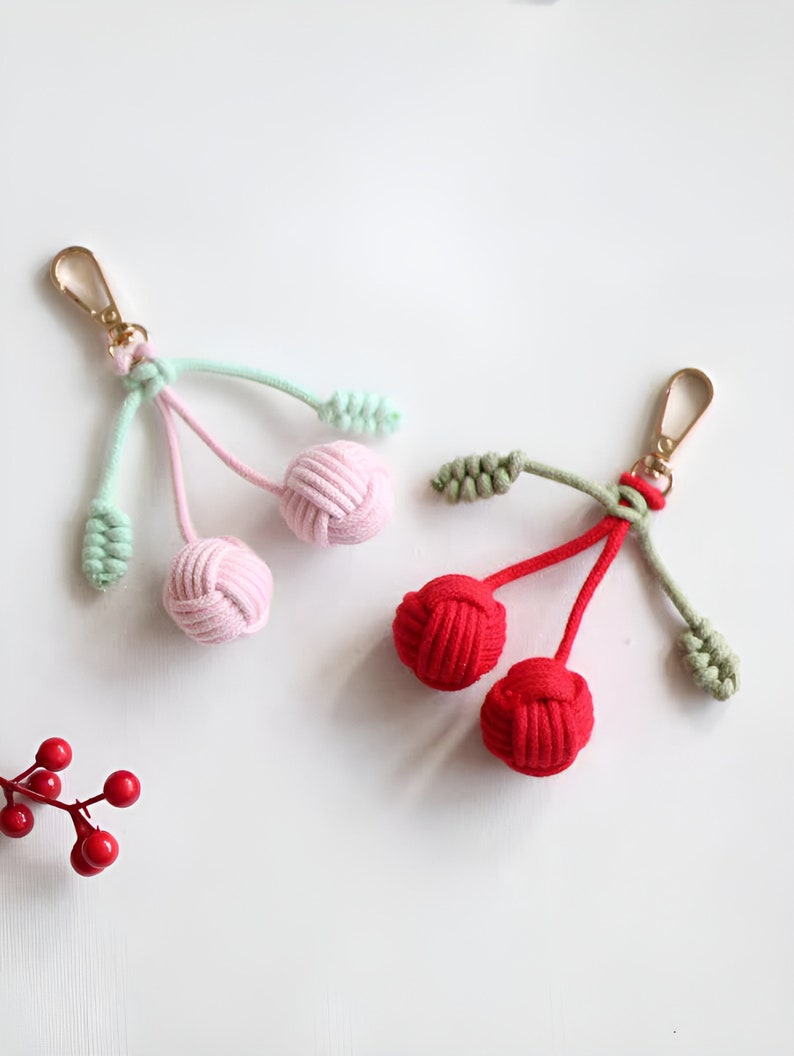 Op de afbeelding: Twee handgeknoopte kers-sleutelhangers. Een hanger is roze met groene bladeren en de andere is rood met groene bladeren.