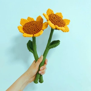 Könnte beinhalten: Zwei gehäkelte Sonnenblumen mit grünen Stielen und Blättern. Die Sonnenblumen sind gelb und braun.